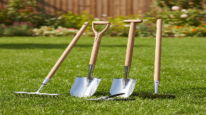 Set of gardening tools including shovels and a rake on grass with flowers in the background